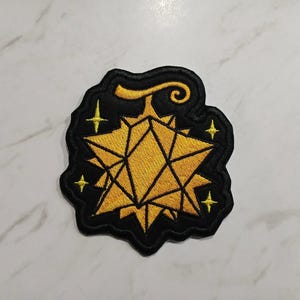 Op de afbeelding: Geborduurde patch met een geometrisch sterontwerp in goud en een zwarte rand. De patch heeft vier kleine gouden sterren en een decoratieve krul. Ideaal voor jassen, tassen of andere stoffen items.