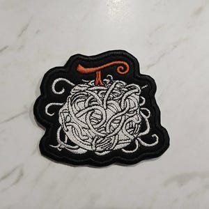May include: Embroidered patch with a white spaghetti design and an orange accent on a black background. The patch has a unique shape with decorative swirls. Suitable for jackets, bags, or other fabric items. Measures approximately 7 cm.