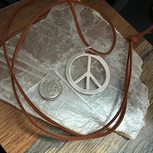 May include: A silver peace sign pendant necklace with a brown leather cord. A US dime is next to the pendant for size comparison. The necklace is displayed on a light gray, textured surface, with a wooden background.