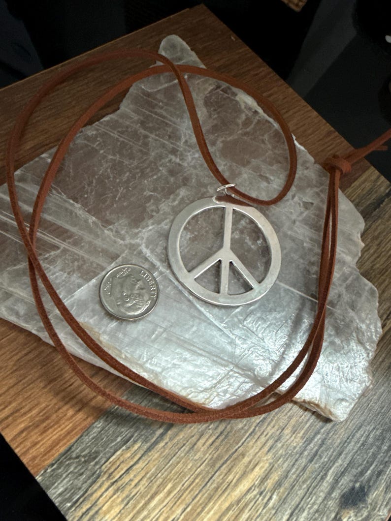 May include: A silver peace sign pendant necklace with a brown leather cord. A US dime is next to the pendant for size comparison. The necklace is displayed on a light gray, textured surface, with a wooden background.