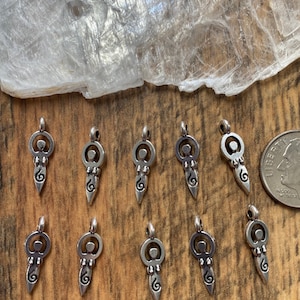 May include: A collection of silver-toned pendants with a stylized female figure design. Each pendant features a circular head, a body with a spiral detail, and a loop for a chain. The pendants are displayed on a wooden surface.