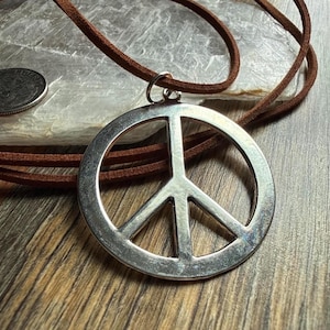 May include: A silver-colored peace sign pendant on a brown leather cord. The pendant is round with a classic peace symbol design. The cord is a thin, natural brown color. The necklace is displayed on a wooden surface.