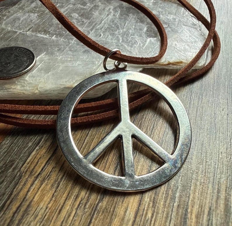 May include: A silver-colored peace sign pendant on a brown leather cord. The pendant is round with a classic peace symbol design. The cord is a thin, natural brown color. The necklace is displayed on a wooden surface.