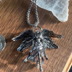 May include: Silver-toned angel pendant necklace. The pendant features an angel with outstretched wings and flowing robes. The necklace has a delicate chain and is displayed on a wooden surface.