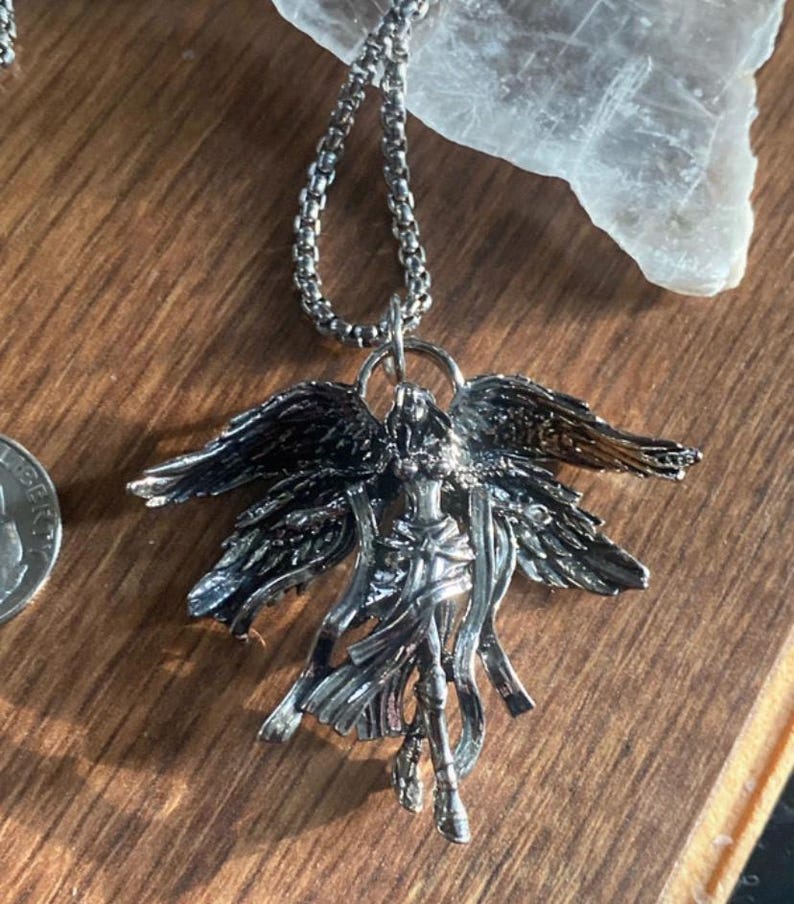 May include: Silver-toned angel pendant necklace. The pendant features an angel with outstretched wings and flowing robes. The necklace has a delicate chain and is displayed on a wooden surface.