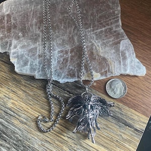 May include: A silver-toned pendant necklace featuring a detailed angel figure with outstretched wings. The pendant hangs from a delicate chain. A U.S. dime is included for size reference. The necklace is displayed on a wooden surface.