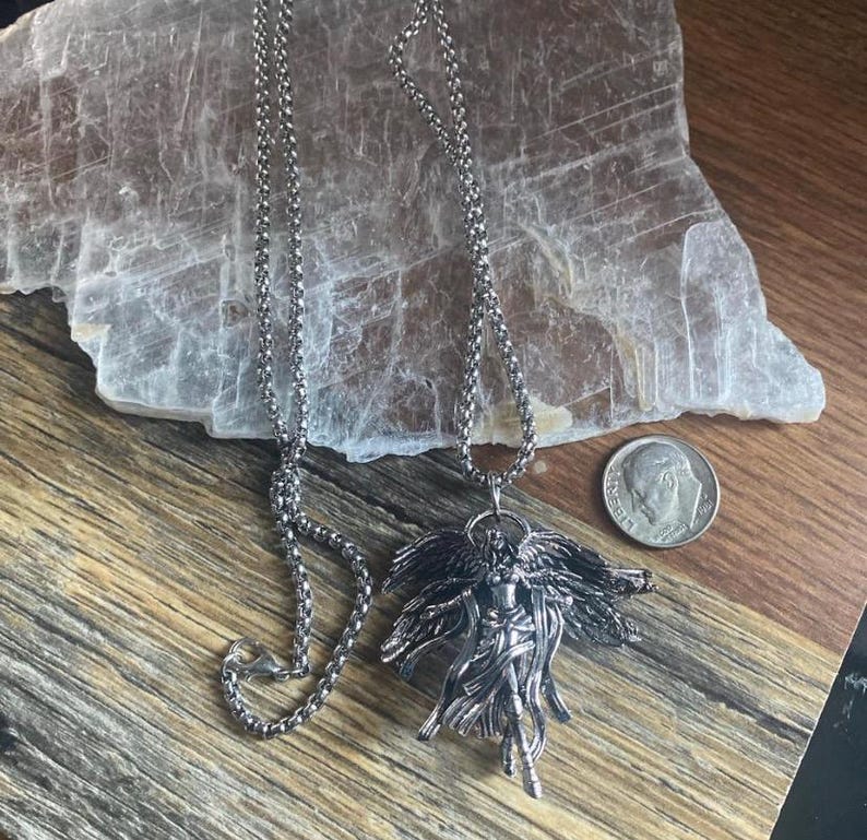 May include: A silver-toned pendant necklace featuring a detailed angel figure with outstretched wings. The pendant hangs from a delicate chain. A U.S. dime is included for size reference. The necklace is displayed on a wooden surface.