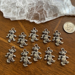 May include: Ten silver-colored dancing bear charms. The bears are standing on their hind legs with their arms raised in the air. They have a smiling face and a furry body.