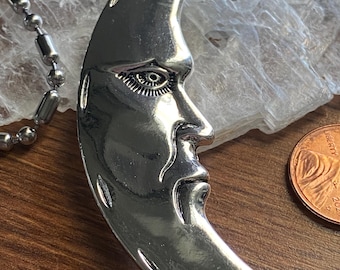 May include: A silver-colored crescent moon pendant with a detailed face and a five-pointed star. The pendant is attached to a silver ball chain. The moon has a detailed face with an eye, nose, and mouth.