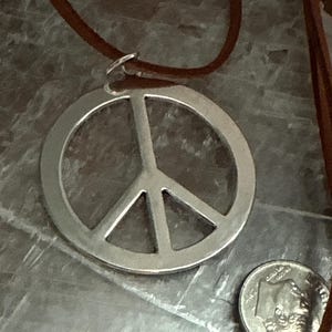 May include: A silver-colored peace sign pendant with a brown leather cord. The pendant is round with a classic peace symbol design. A dime is next to the pendant for size comparison. The cord is attached to the pendant with a small silver ring.