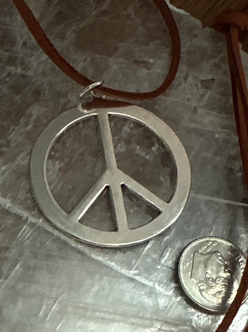 May include: A silver-colored peace sign pendant with a brown leather cord. The pendant is round with a classic peace symbol design. A dime is next to the pendant for size comparison. The cord is attached to the pendant with a small silver ring.