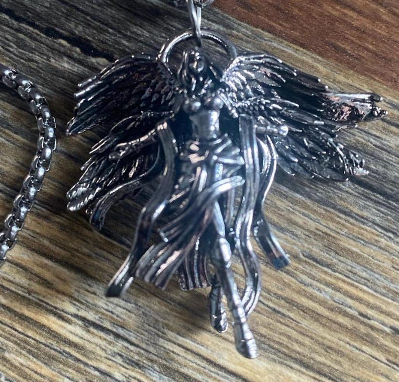 May include: A silver-toned pendant necklace featuring an angel figure with outstretched wings and flowing robes. The detailed pendant is suspended from a matching chain, creating a gothic or fantasy aesthetic. The necklace is displayed on a wooden surface.