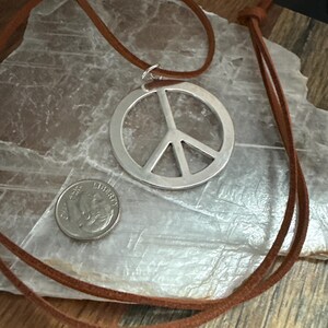 May include: A silver peace sign pendant necklace with a brown leather cord. A U.S. dime is next to the pendant for size comparison. The necklace is displayed on a light-colored, textured surface.