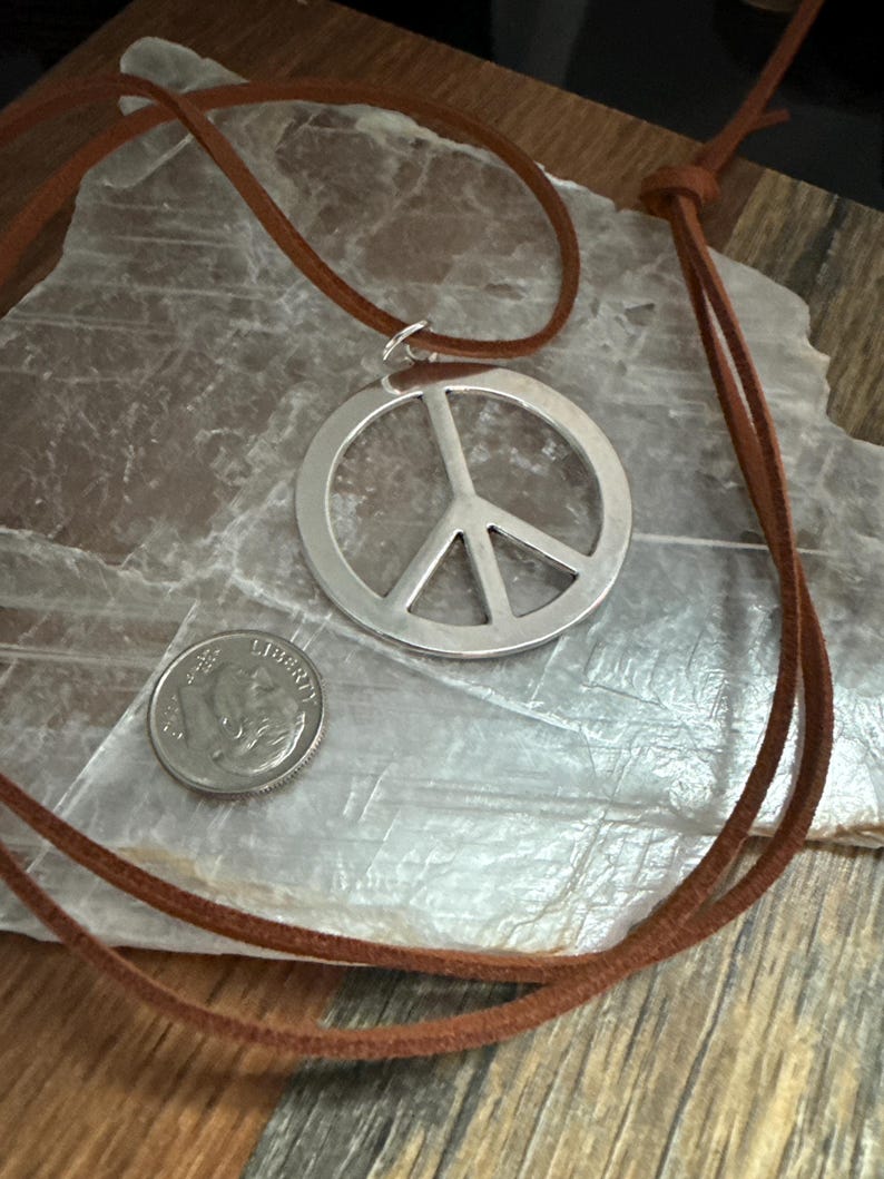 May include: A silver peace sign pendant necklace with a brown leather cord. A U.S. dime is next to the pendant for size comparison. The necklace is displayed on a light-colored, textured surface.