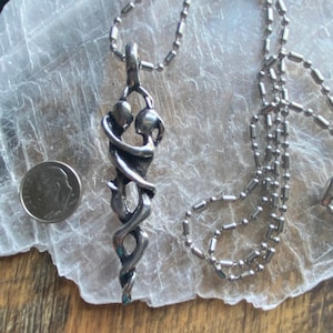 May include: A silver necklace with a pendant depicting two figures intertwined in an abstract design. The pendant is about the size of a US quarter.