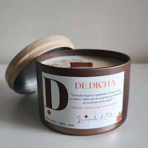 May include: A brown metal candle tin with a wooden lid, featuring a white label with text and a large letter "D". The candle has a wooden wick and a light-colored wax. The label includes the words "DE DICHA".