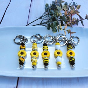 May include: Five sunflower keyrings with different bead patterns. Each keyring has a silver key ring and a silver bead at the bottom. The beads are yellow, black, and white.