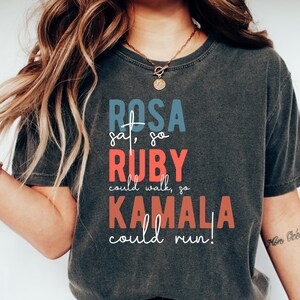 May include: A dark grey t-shirt with a graphic print that reads "ROSA sat, so RUBY could walk, so KAMALA could run!"