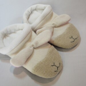 May include: A pair of white and beige baby slippers with pink ears and a stitched face. The slippers are made of soft, plush fabric.