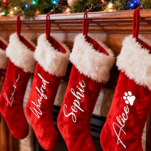 Puede incluir: Cinco calcetines navideños rojos con ribete de piel sintética blanca cuelgan de una repisa de madera. Cada calcetín tiene un nombre en escritura blanca: Sophie, Laura, Winifred, Sophie y Alice. El último calcetín tiene una huella de pata blanca.