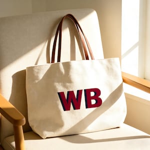 Pode incluir: Uma sacola tote creme com alças de couro marrom e as letras "WB" em vermelho e azul marinho. A sacola está em uma cadeira bege, perto de uma janela.