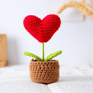 May include: A crocheted flower with a red heart-shaped bloom, green leaves, and a brown pot. The stem is green. The flower is sitting on a book.