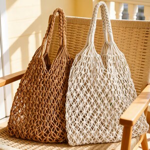 May include: Two macrame tote bags, one in a warm brown and the other in a natural off-white, rest on a woven chair. The bags have braided handles and open, netted bodies, ideal for carrying groceries or beach essentials. The chair is made of light-coloured wood.