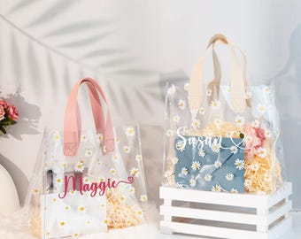 May include: Two clear plastic tote bags with daisy print and personalized names. The bag on the left has pink handles and says "Maggie" in pink script. The bag on the right has beige handles and says "Susan" in white script.