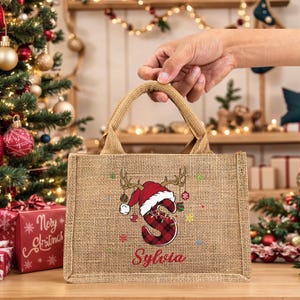 May include: A burlap tote bag with a Christmas theme. The bag features a red and black plaid letter "S" wearing a Santa hat and antlers, with the name "Sylvia" below. The bag has a natural tan color and a woven texture.