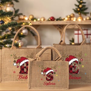 May include: Three burlap tote bags with handles, each featuring a Christmas design. The design includes a Santa hat, antlers, and a letter in a plaid pattern. Names are printed below the letters.