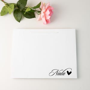 May include: A white gift bag with a handle, featuring the name "Nala" in elegant script with a black heart. A pink rose and green leaves are placed on the top left corner of the bag.