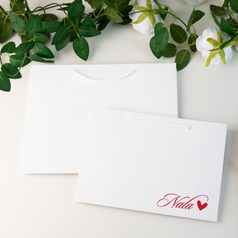May include: Two white gift bags with white rope handles. One bag has the name "Nala" in red script with a red heart. Green leaves and white flowers are in the background.