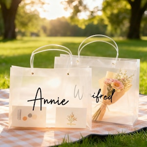 Op de afbeelding: Twee doorzichtige plastic cadeauzakken met witte handvatten. Eén tas heeft de naam "Annie" in zwart schrift, de andere "Wifred" in grijs schrift. Eén tas bevat een boeket roze en gele bloemen. Beide tassen hebben kleine kaarten.