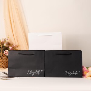 May include: Three gift bags in the image. Two black bags with the name "Elizabeth" in white script and a heart symbol. A white bag is behind the black bags. All bags have handles. The background includes flowers and a sheer curtain.