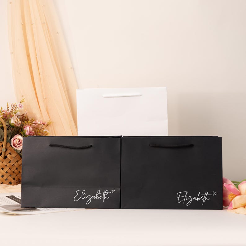 May include: Three gift bags in the image. Two black bags with the name "Elizabeth" in white script and a heart symbol. A white bag is behind the black bags. All bags have handles. The background includes flowers and a sheer curtain.