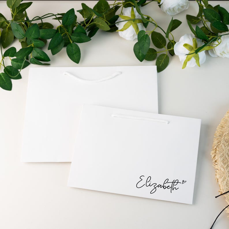 May include: Two white gift bags with white rope handles. One bag has the name "Elizabeth" written in cursive with a small heart. Green leaves and white flowers are in the background.