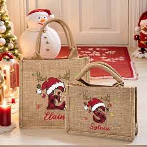 May include: Two burlap tote bags with brown handles. Each bag features a Christmas design with a red plaid letter, antlers, and a Santa hat. The larger bag says "Elain" and the smaller bag says "Sylvia".