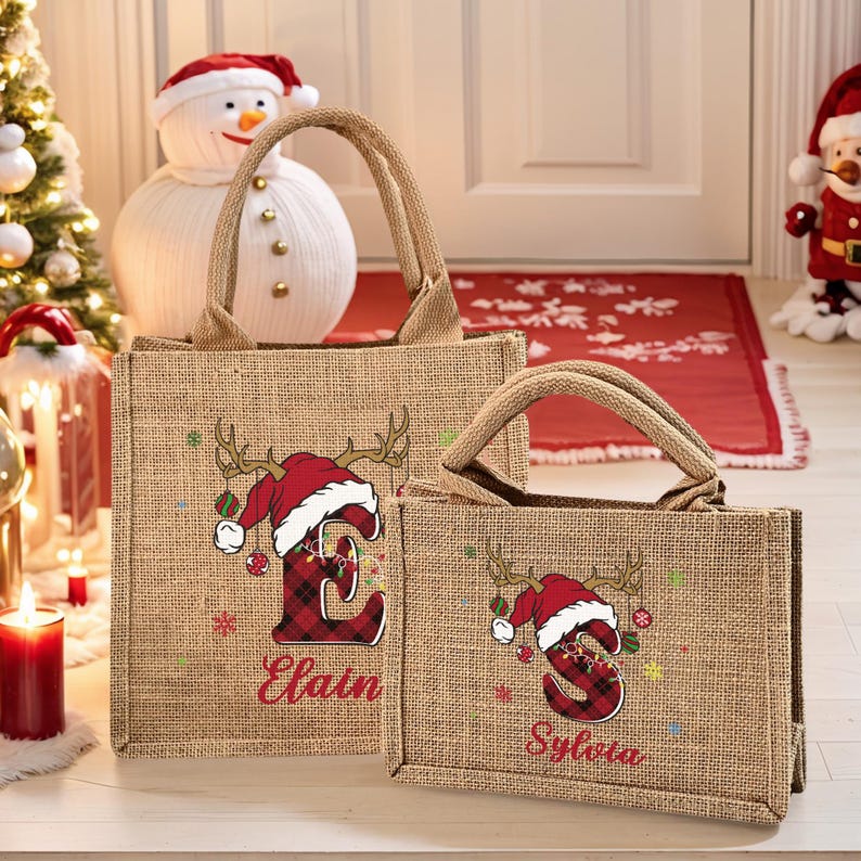 May include: Two burlap tote bags with brown handles. Each bag features a Christmas design with a red plaid letter, antlers, and a Santa hat. The larger bag says "Elain" and the smaller bag says "Sylvia".