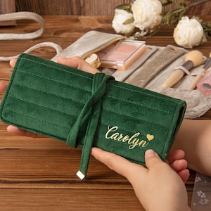 Puede incluir: Un estuche enrollable para brochas de maquillaje de terciopelo verde oscuro con el nombre "Carolyn" y un corazón dorado bordado en la parte delantera. El rollo está atado con una cinta de terciopelo a juego. Contiene brochas y cosméticos.
