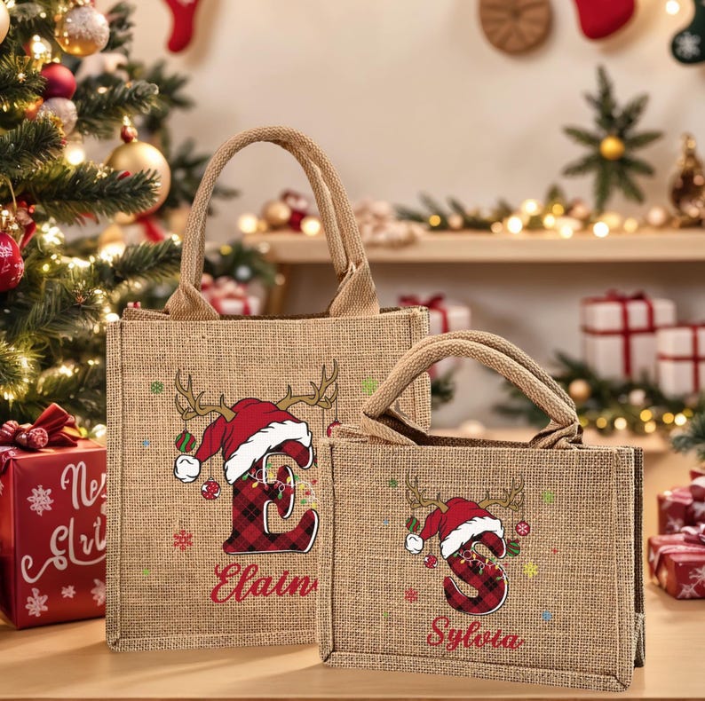 May include: Two burlap tote bags with Christmas-themed designs. Each bag features a reindeer antler and Santa hat design with a letter and name. The larger bag says "Elain" and the smaller bag says "Sylvia".