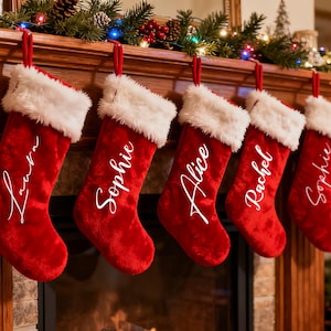 May include: Five red Christmas stockings with white faux fur trim hang from a wooden mantelpiece. Each stocking has a name in white script: Laura, Sophie, Alice, Rachel, and Sophie. The mantelpiece is decorated with greenery and lights.
