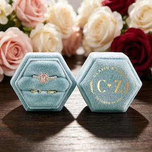 May include: A light blue velvet hexagonal ring box with two slots. The left side holds a silver ring with a pink rectangular stone and a gold ring with a blue stone. The right side features gold text: CLAVIN & ZOE, C+Z, OCTOBER 11, 2025.