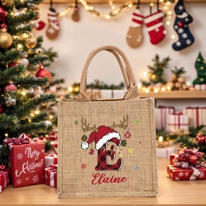 May include: A burlap tote bag with a Christmas theme. The bag features a red and black plaid letter "E" wearing a Santa hat and reindeer antlers. The name "Elaine" is printed below the letter. Christmas decorations are in the background.