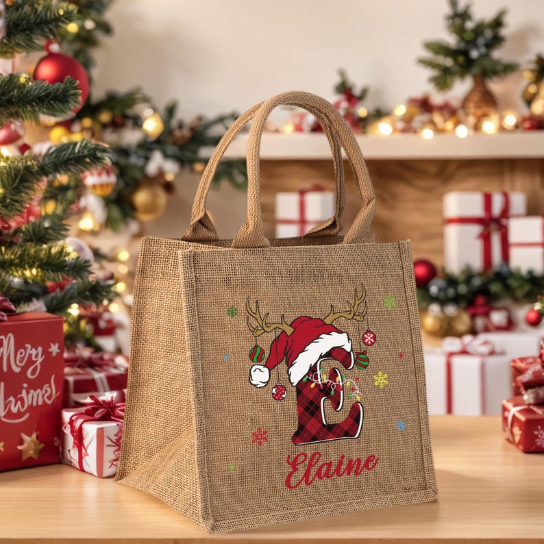 May include: A burlap tote bag with a Christmas-themed design. The bag features a red and black plaid letter "E" wearing a Santa hat with antlers and the name "Elaine" in red. Colorful ornaments and snowflakes decorate the bag.