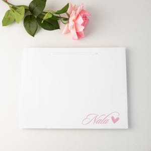 May include: A white gift bag with a thin white handle and the name "Nala" in pink script with a pink heart. A pink rose and green leaves are in the background.