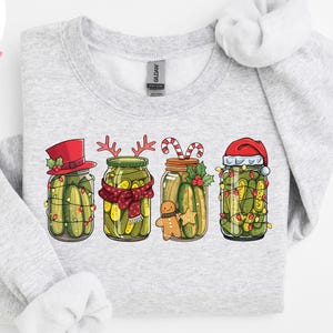 Peut inclure: Sweat-shirt gris clair avec une illustration de quatre bocaux de cornichons décorés pour Noël. Chaque bocal a un design festif unique, avec un bonnet de Père Noël, des bois de renne et des cannes en sucre. Le mot "ASH" est imprimé en bas.
