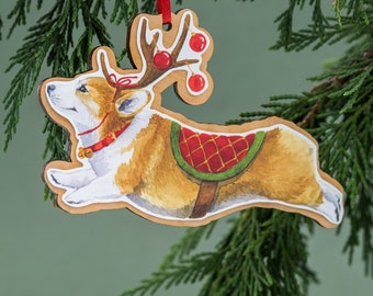 May include: A wooden Christmas bauble featuring a cartoon illustration of a corgi dog wearing a red and green reindeer costume with red and green holly berries. The corgi is jumping in the air with its tongue sticking out.