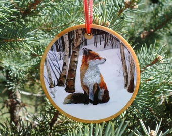 Peut inclure: Une décoration ronde en bois avec une image peinte d'un renard roux dans un décor de forêt enneigée. Le renard est assis et regarde vers la droite. La décoration est munie d'un ruban rouge.