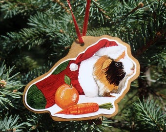Puede incluir: Adorno navideño de madera con un conejillo de indias con gorro de Papá Noel y un jersey rojo. También se representan una naranja y una zanahoria. El adorno cuelga de una cinta roja.