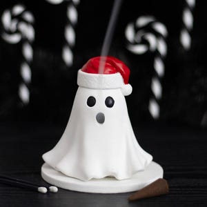 May include: A white ghost-shaped incense burner wearing a red and white Santa hat. The ghost has two black circular eyes and an open circular mouth. A thin trail of smoke rises from the top of the hat. The figure sits on a circular white base.